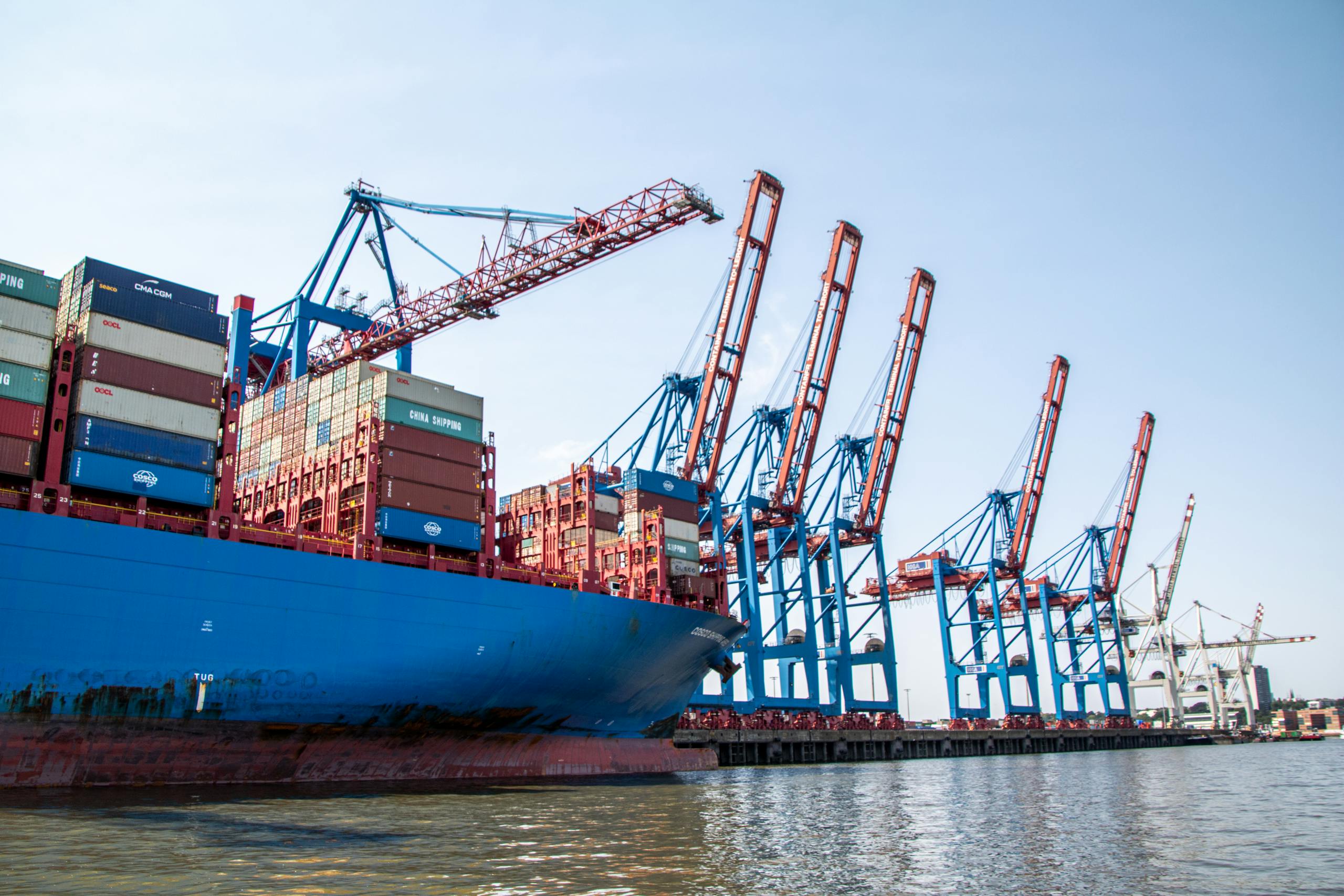 A large container ship docked at the Port of Hamburg, surrounded by red and blue cranes.